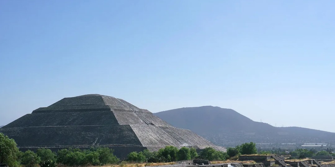 La pirámide del Sol en Teotihuacan al frente con un cerro y el cielo despejado detrás.