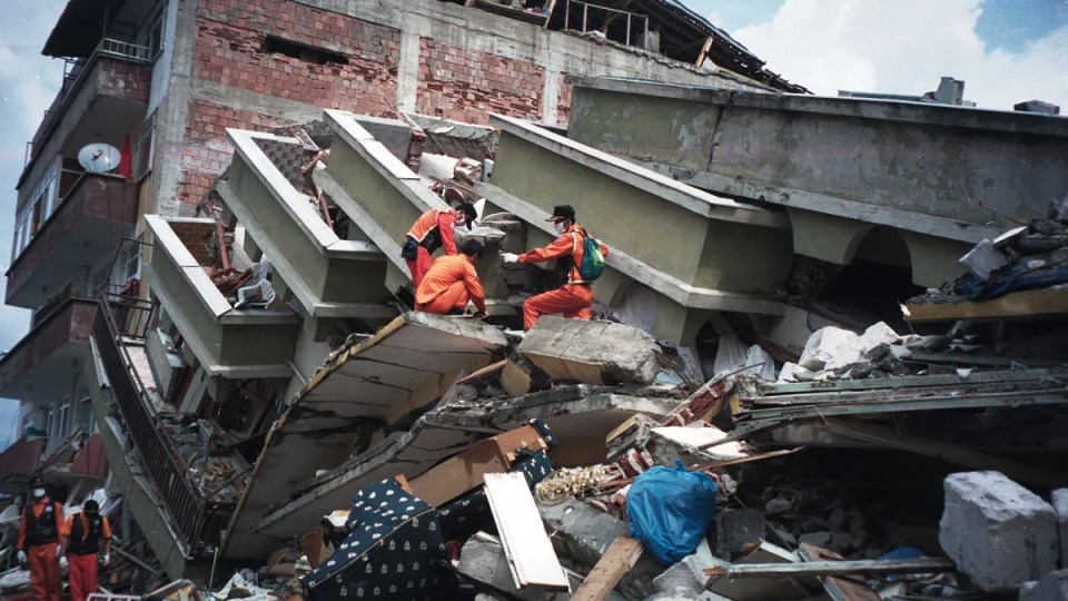 Tres rescatistas, vestidos en color naranja, buscan entre los escombros de un edificio caído. Detrás del edificio caído hay otro edificio de ladrillos rojos, el cual aún se encuentra de pie, pero tiene algunas paredes dañadas