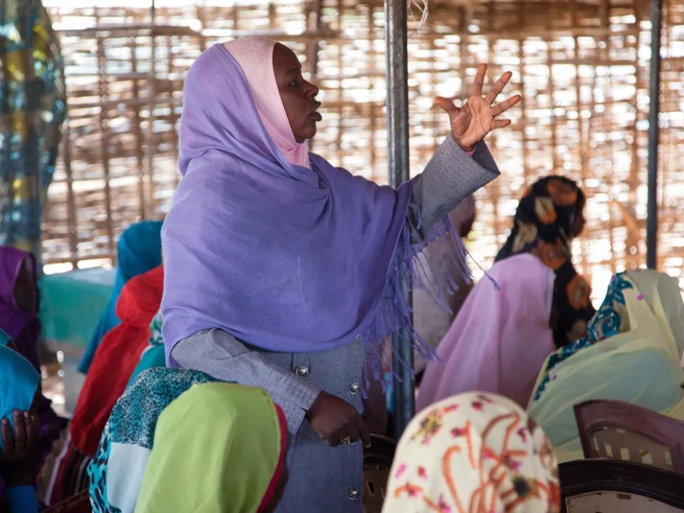 Al centro una mujer con un hijab lila está de pie para hablar en una junta. A su alrededor hay otras mujeres sentadas.
