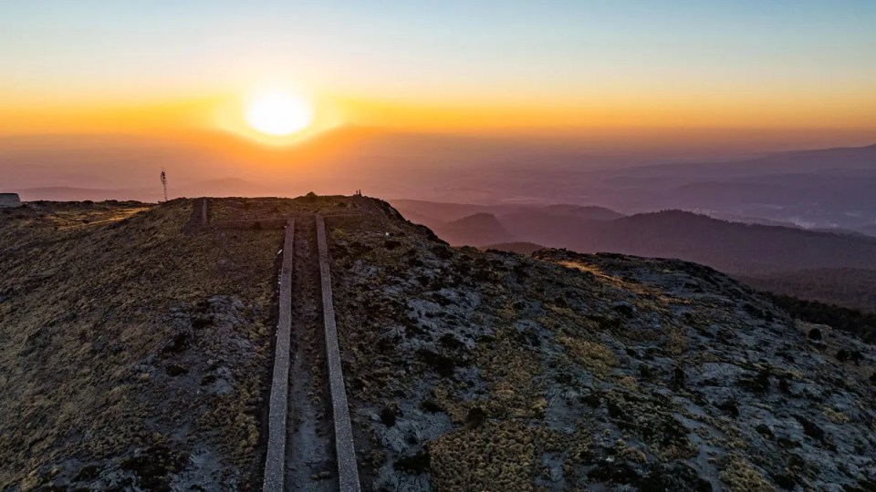 Imagen aérea del amanecer en el Monte Tláloc. Al fondo se ve el Sol (en tonos naranjas) saliendo entre nubes y al frente la punta del Monte Tláloc con una calzada alzándose al centro.