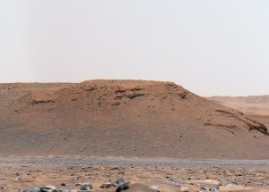 Una foto de una colina roja con una cima plana al fondo con rocas sobre en un paisaje al frente. 